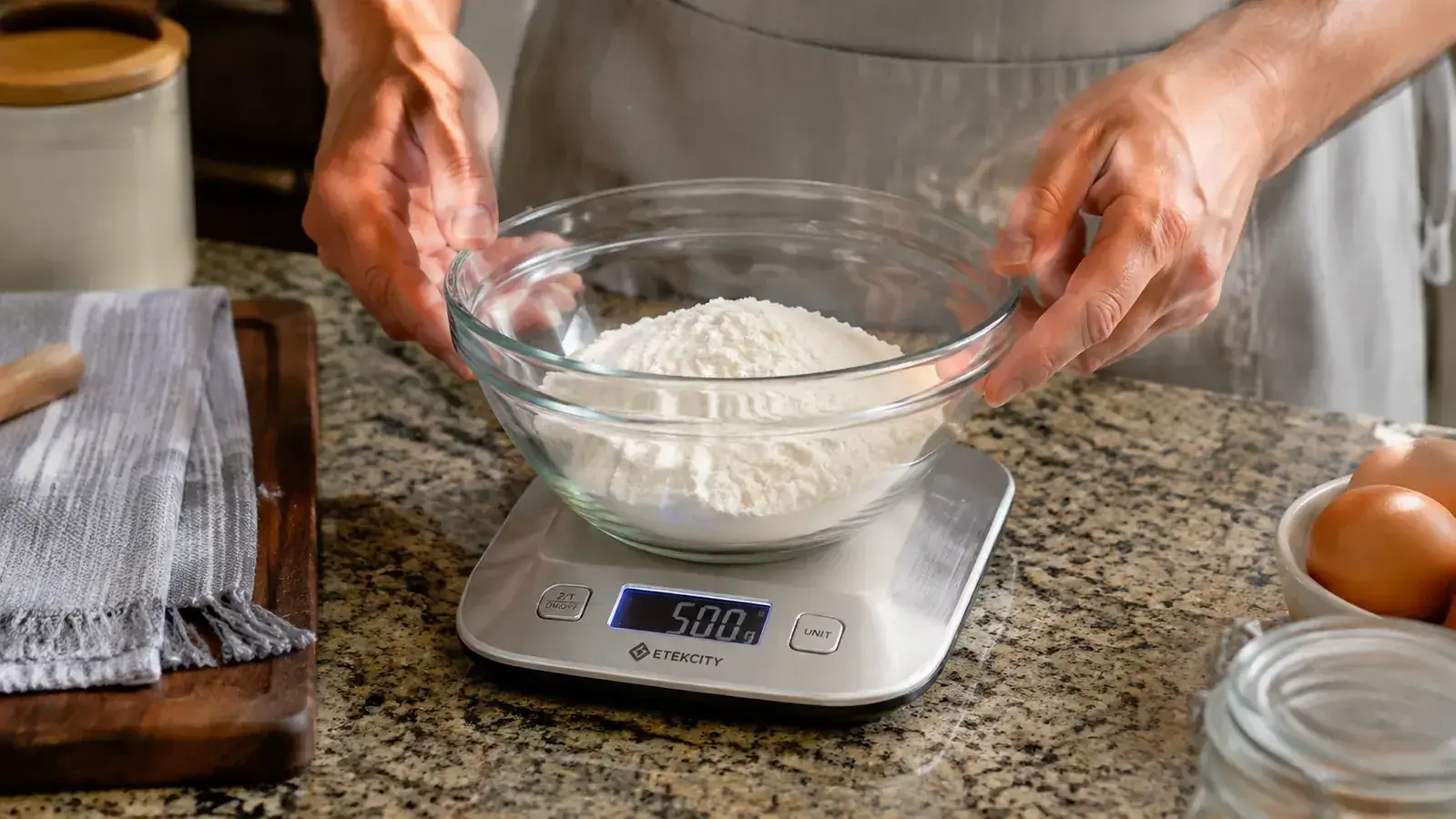 Hand placing a mixing bowl of flour onto a stainless steel digital kitchen scale showing a weight reading on the display
