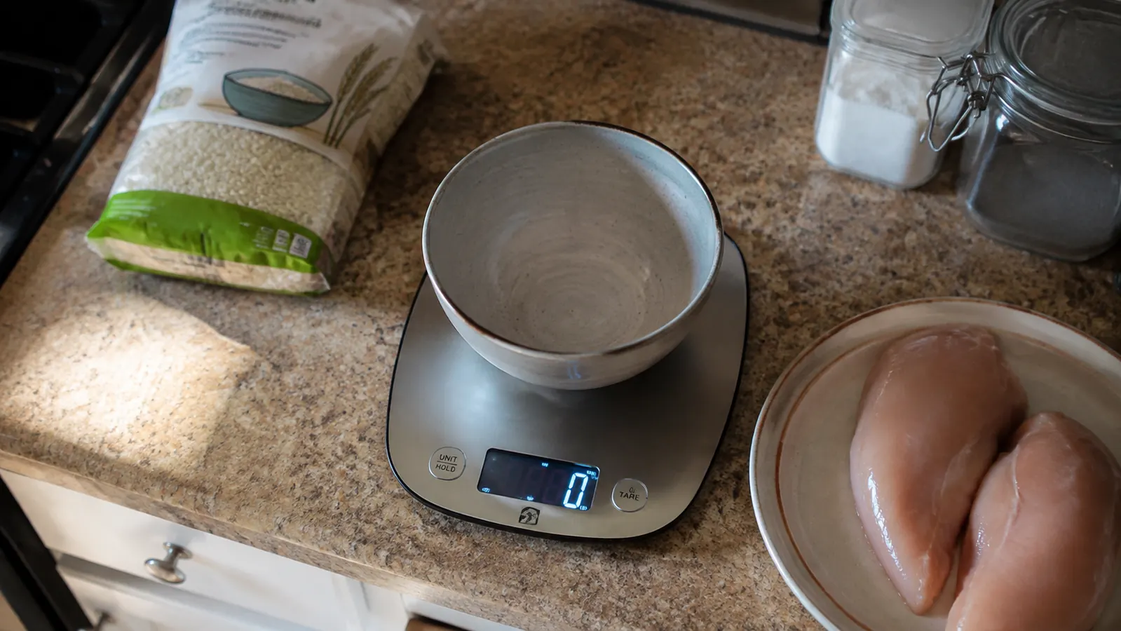 A home kitchen counter with a stainless steel digital kitchen scale, a mixing bowl, and containers ready for portioning a meal