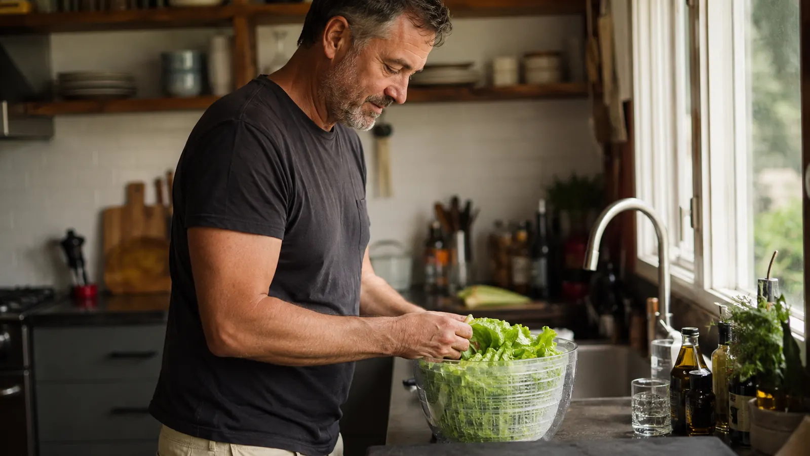 Home cook standing at a kitchen counter next to a glass salad spinner full of freshly washed lettuce